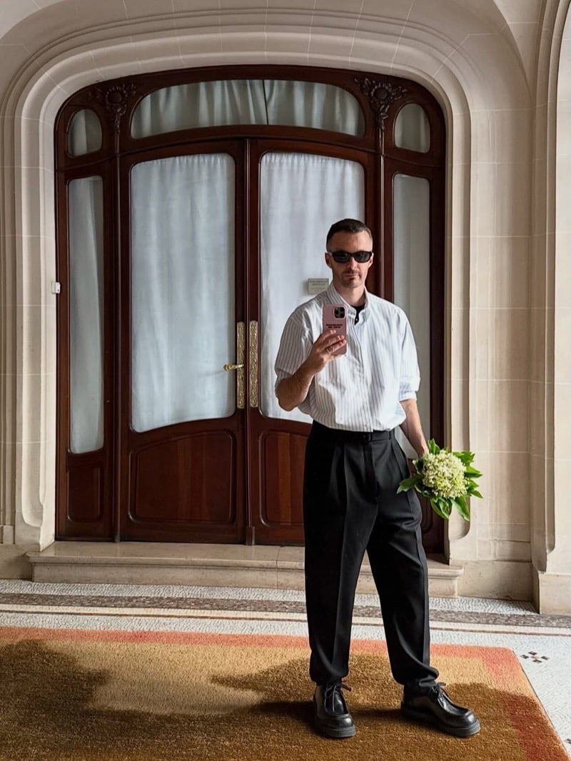 Kris Van Assche takes a mirror selfie in sunglasses, a striped shirt, and pleated trousers, holding flowers