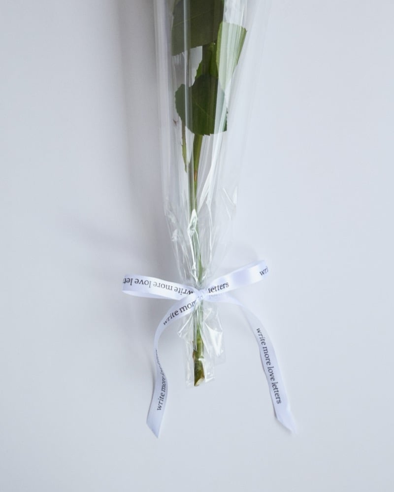 A single long-stemmed rose wrapped in cellophane and tied with a ribbon reading write more love letters