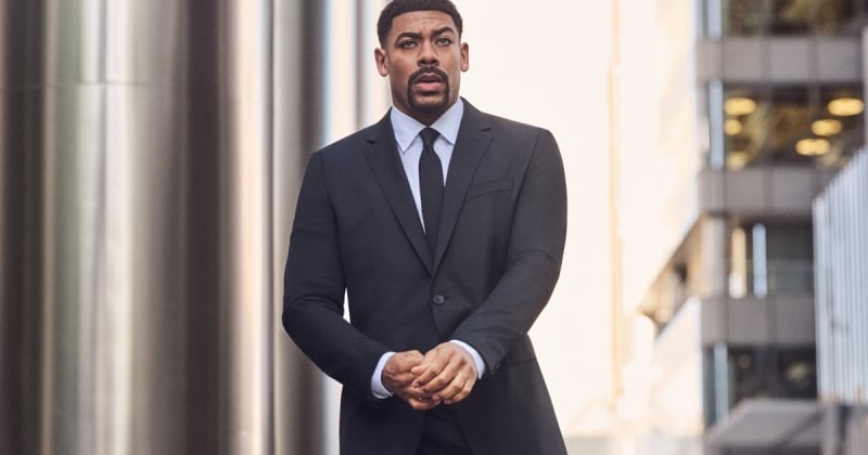 Aaron Pierre wears a black suit with a white shirt and black tie in front of a city building for BOSS's fall-winter 2026 campaign