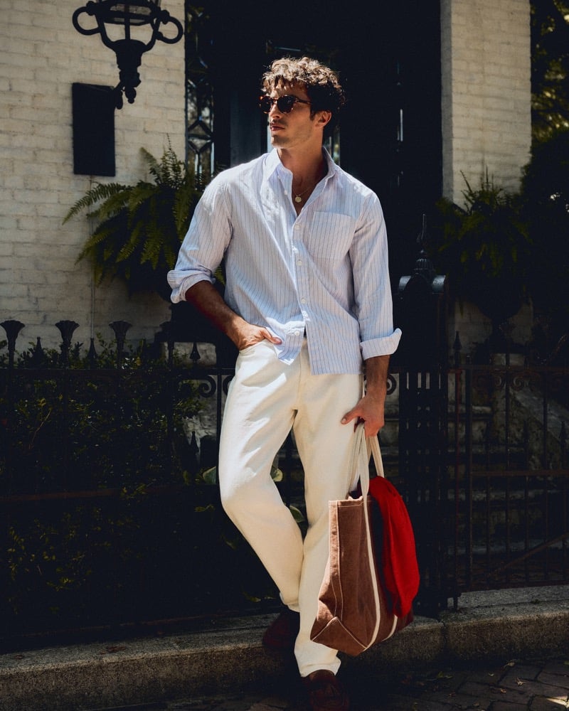 Raphael Diogo in a striped poplin shirt and white jeans carrying a suede tote outside a brick townhouse