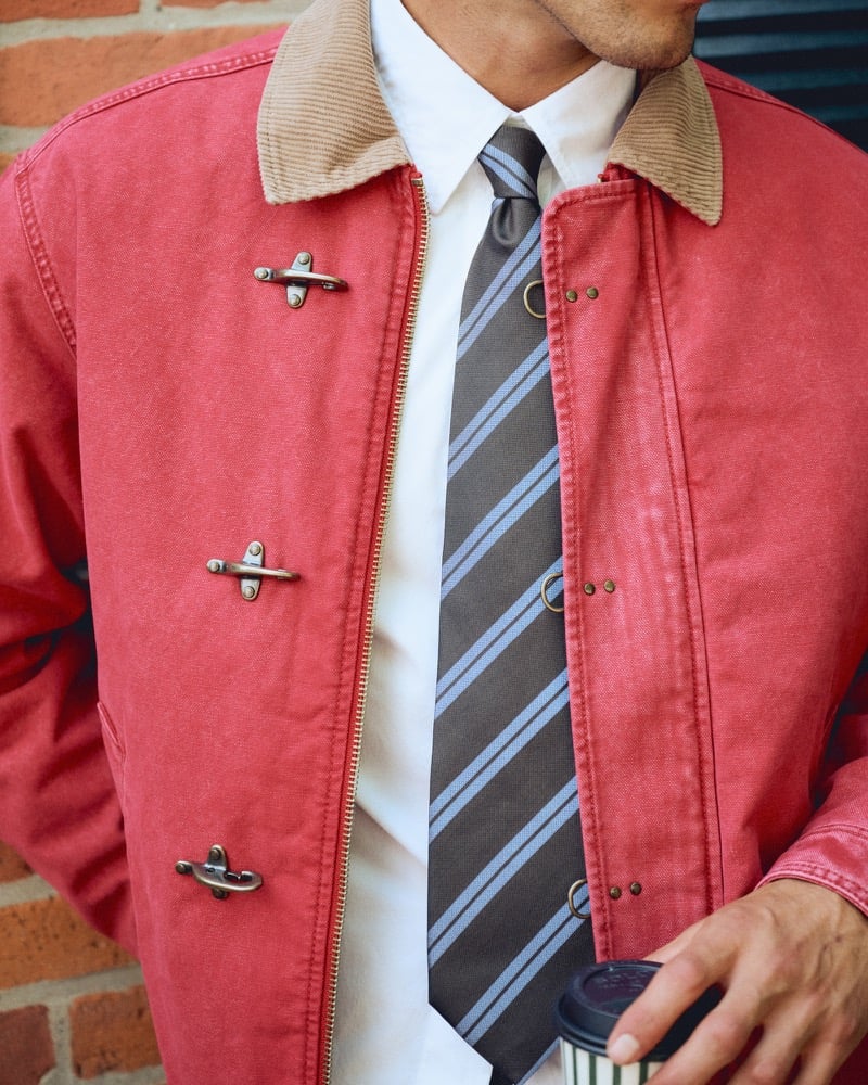 Close-up of a red canvas jacket with fireman clasps and a striped tie, coffee cup in hand