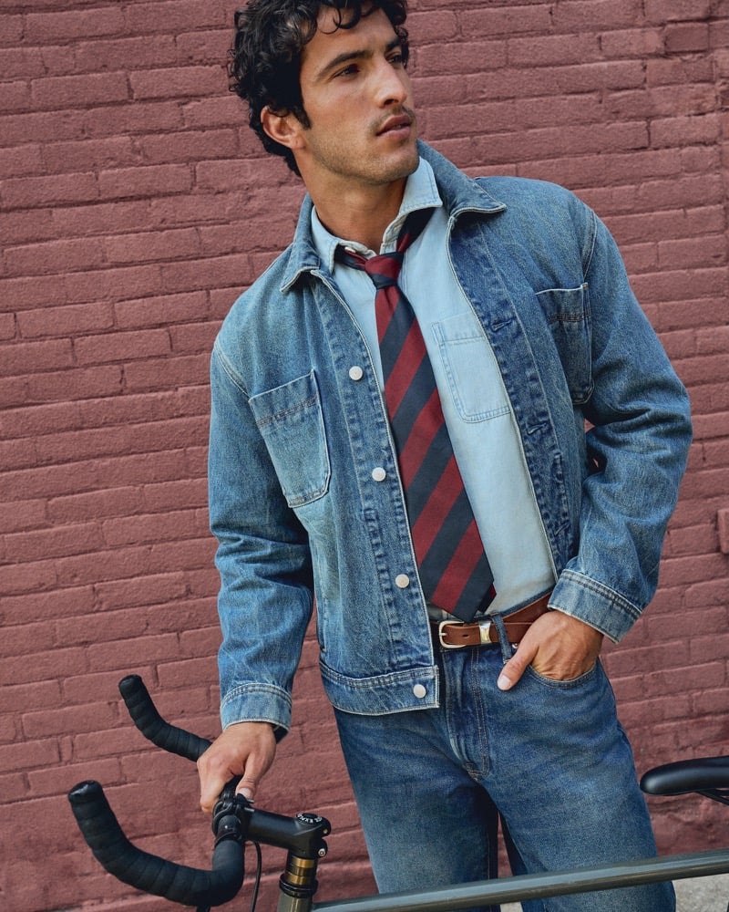 Raphael Diogo in a denim trucker jacket over a chambray shirt with a striped tie, gripping a bicycle handlebar