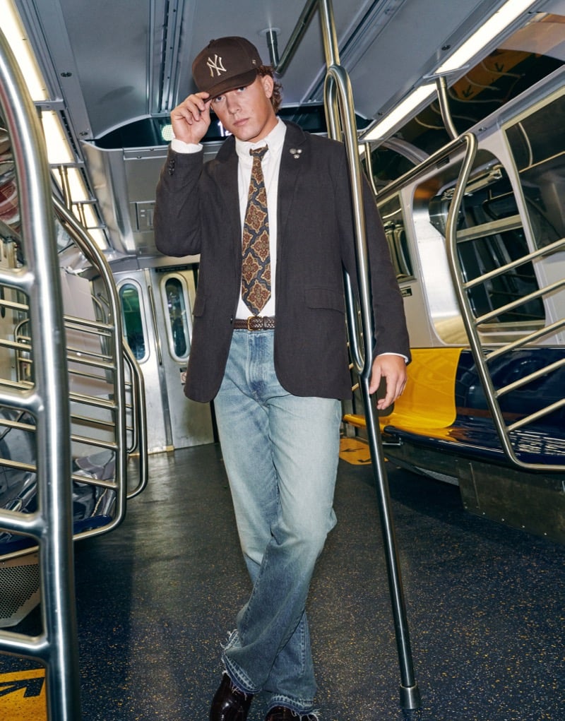 Jaxson Dart adjusts his New York Yankees cap while walking through a subway car in a dark pinstripe blazer, patterned tie, and straight-leg jeans