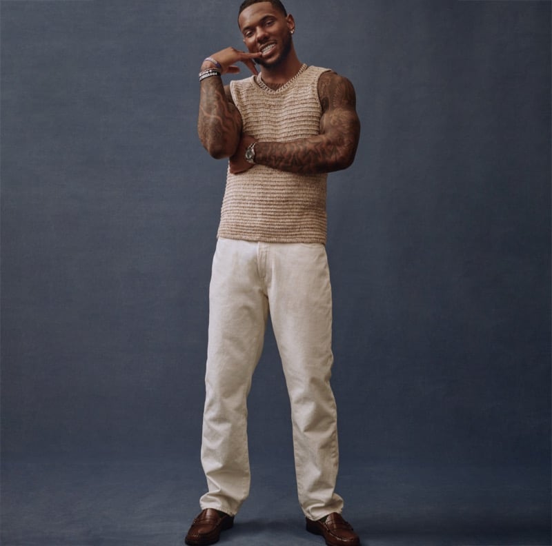 Malik Nabers stands against a studio backdrop in a sleeveless knit sweater and relaxed trousers, tattoos visible on both arms