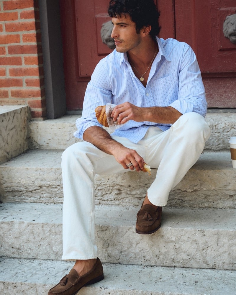 Raphael Diogo seated on stone steps in a striped poplin shirt, white jeans, and brown suede tassel loafers, eating a croissant