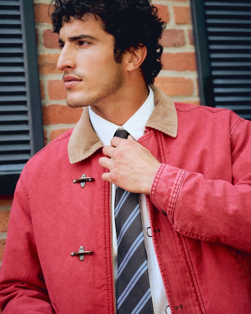 Close-up of Raphael Diogo adjusting a gray striped tie beneath a red corduroy-collar jacket