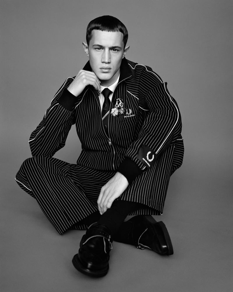 Achille Genet sits in a pinstripe zip-up track jacket and matching trousers with a tie and laurel-wreath badges for Fred Perry x Kris Van Assche, photographed in black and white