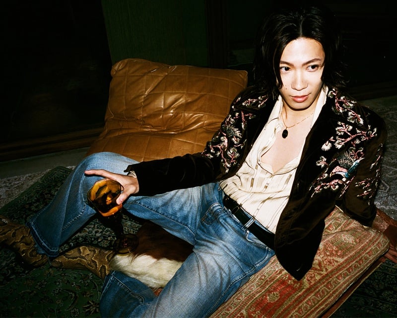 Yuta Jinguji sits on the floor with a drink in a floral-embroidered velvet jacket and flared jeans for AMIRI's fall-winter 2026 campaign