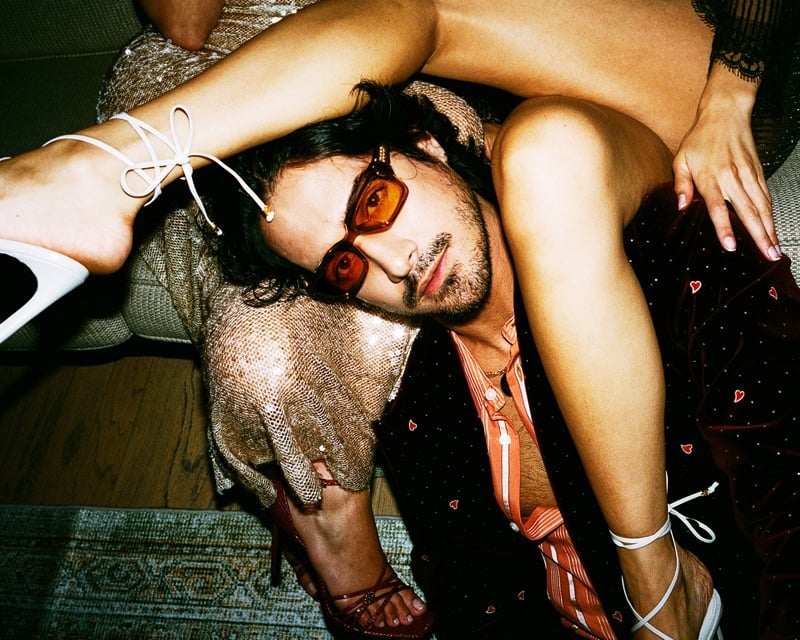 Avan Jogia lies on the floor in amber sunglasses and a heart-embroidered velvet suit as a partygoer's legs rest over him for AMIRI's fall-winter 2026 campaign