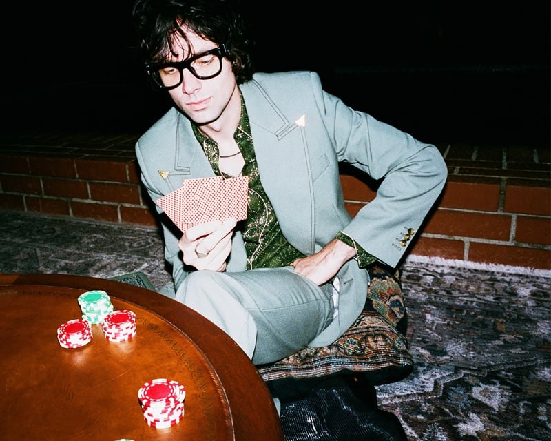Chandler Lucy of The Hellp holds a hand of cards in a sage tailored jacket over a metallic shirt at a poker table for AMIRI's fall-winter 2026 campaign