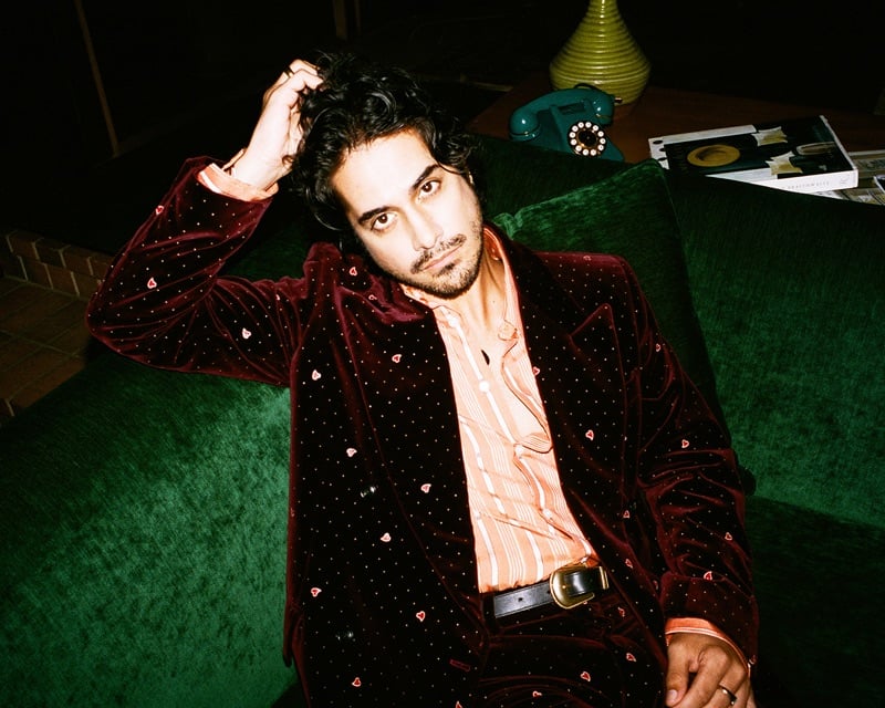 Avan Jogia reclines on a green velvet sofa in a heart-embroidered burgundy velvet suit and striped shirt for AMIRI's fall-winter 2026 campaign
