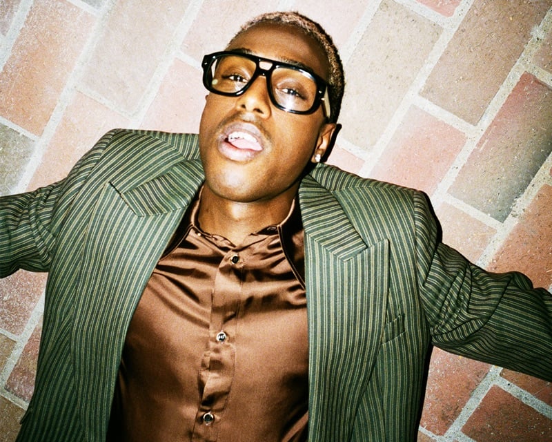 Bakar lies on brick paving in an olive pinstripe jacket over a brown satin shirt for AMIRI's fall-winter 2026 campaign