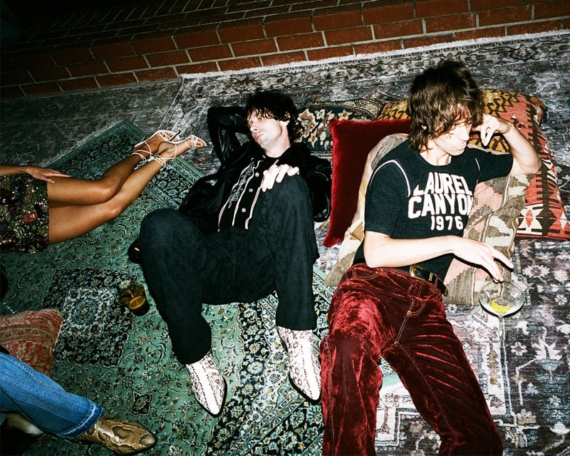 The Hellp sprawl across layered rugs poolside, one in a leather jacket and python boots, the other in a Laurel Canyon tee and burgundy velvet trousers, for AMIRI's fall-winter 2026 campaign