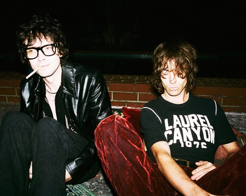 Chandler Lucy and Noah Dillon of The Hellp sit poolside at night, one in a leather jacket with a cigarette, the other in a Laurel Canyon knit and burgundy velvet trousers, for AMIRI's fall-winter 2026 campaign