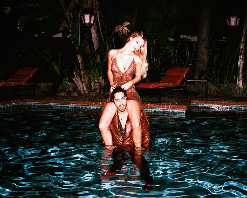 Avan Jogia wades through a lit pool at night with Bella Podaras on his shoulders in a lace slip dress for AMIRI's fall-winter 2026 campaign