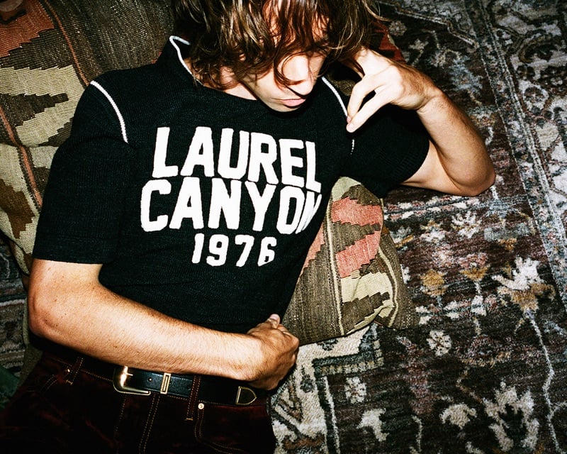 Noah Dillon of The Hellp lies against a kilim cushion in a Laurel Canyon 1976 knit and burgundy velvet trousers for AMIRI's fall-winter 2026 campaign