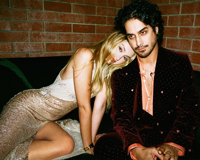 Avan Jogia sits with Bella Podaras against a brick wall in a heart-embroidered burgundy velvet suit for AMIRI's fall-winter 2026 campaign