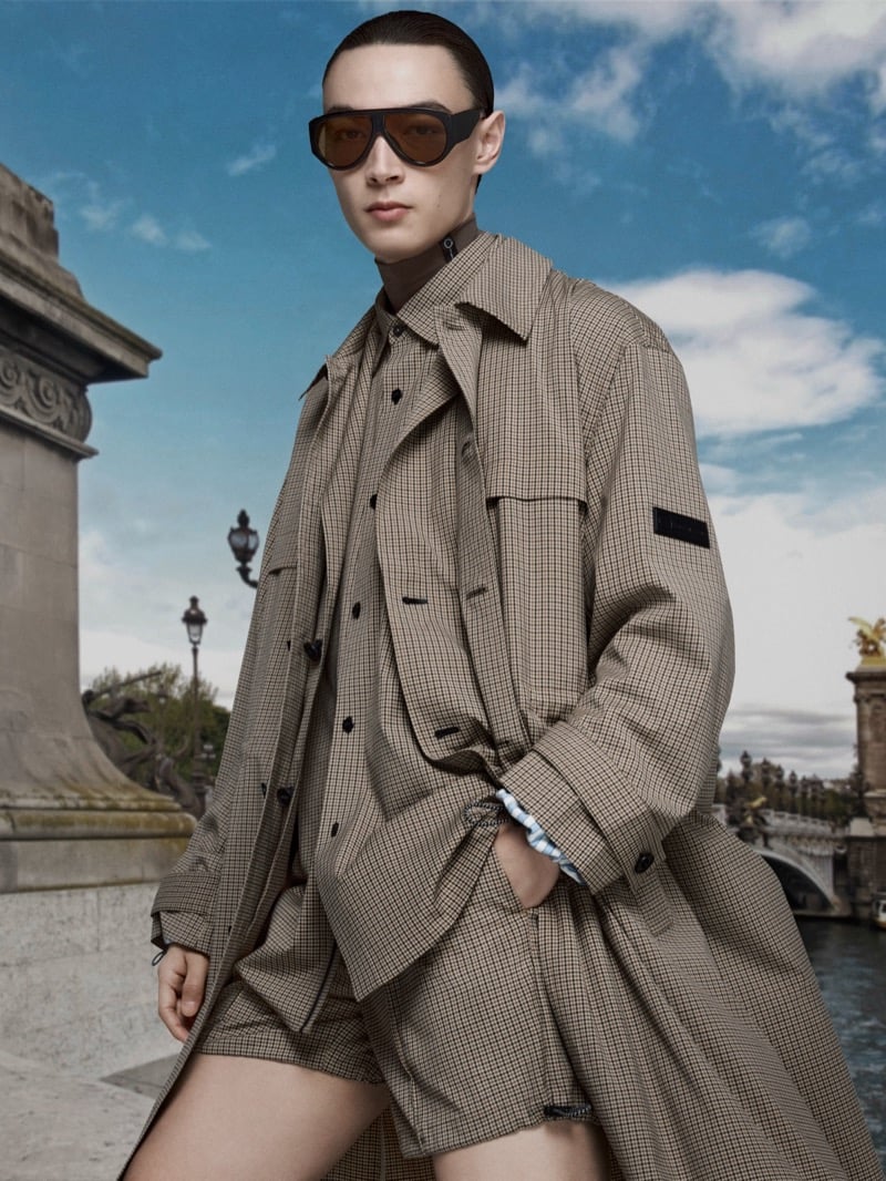 Pascal Thulin wears a checked trench coat over a matching shirt and shorts with aviator sunglasses beside a Paris bridge for ANTAZERO season two