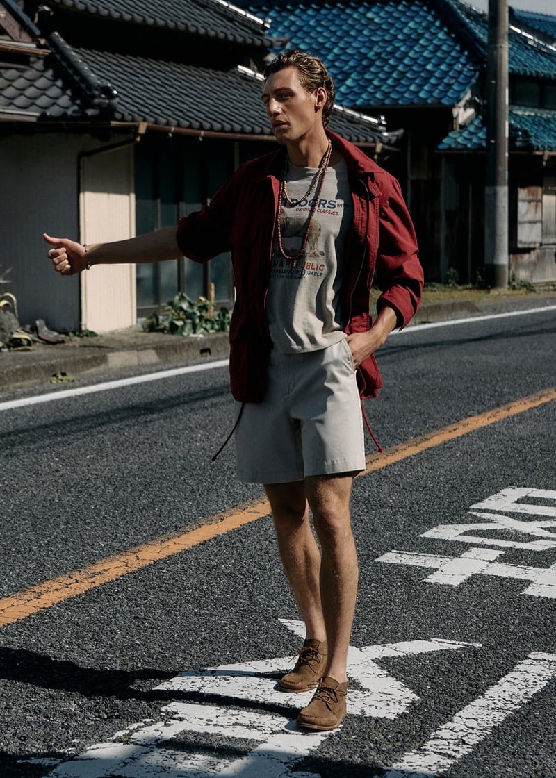 Aubrey O'Mahony hitchhiking on a Japanese street in a burgundy archive reissue field jacket, graphic T-shirt, and shorts with suede boots