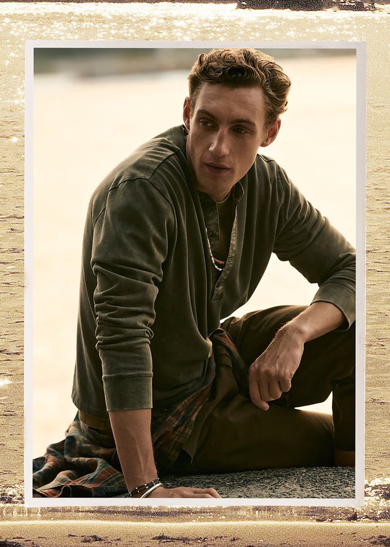 Aubrey O'Mahony seated on a rock in an olive archive reissue henley with a plaid shirt tied at his waist