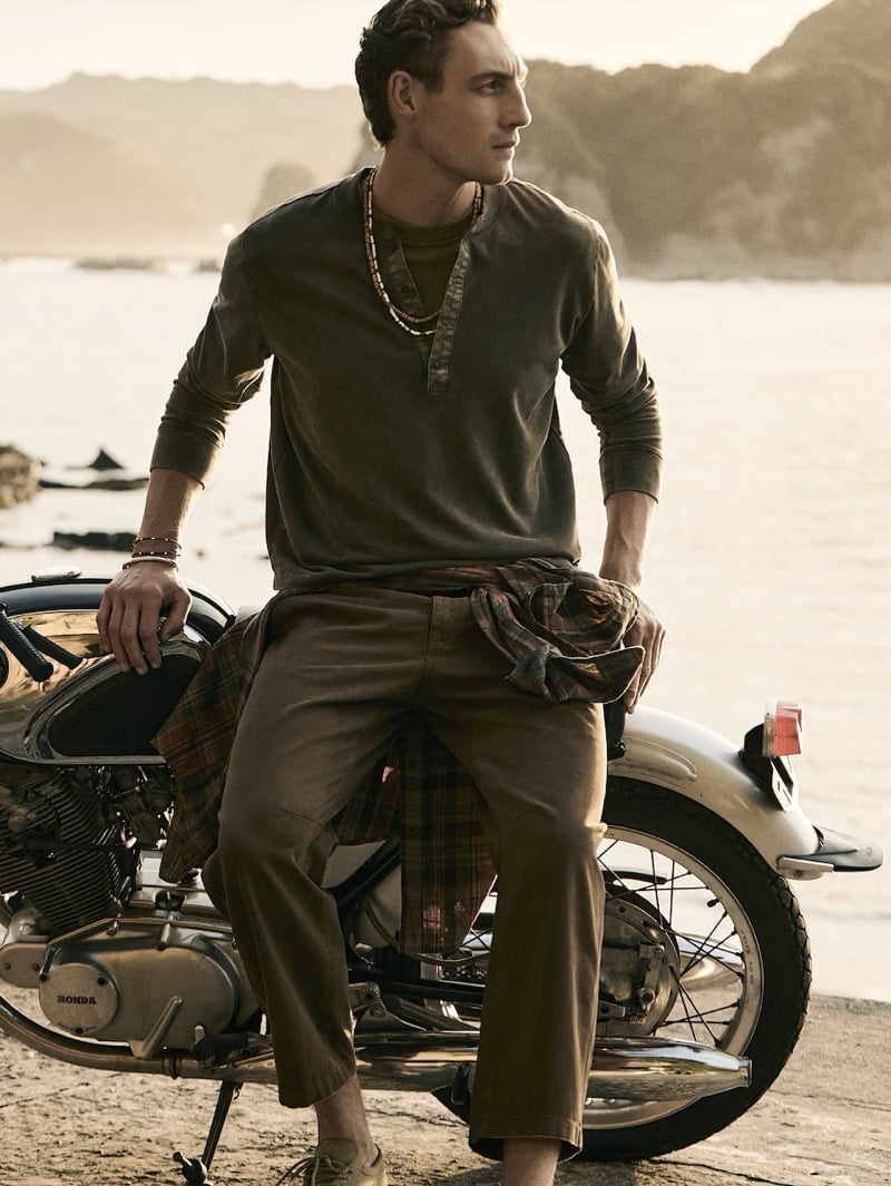 Aubrey O'Mahony seated on a vintage Honda motorcycle in an olive henley and relaxed tapered chinos with a plaid shirt tied at the waist
