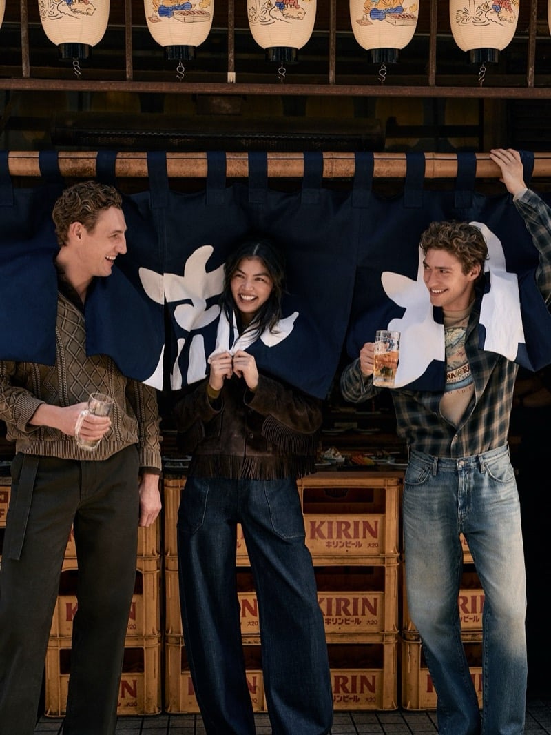 Aubrey O'Mahony in a brown cable-knit vest, Rina Fukushi in a suede fringe jacket, and Valentin Humbroich in a plaid shirt over a graphic tee share drinks beneath a shop's noren curtain for Banana Republic's September 2026 edit