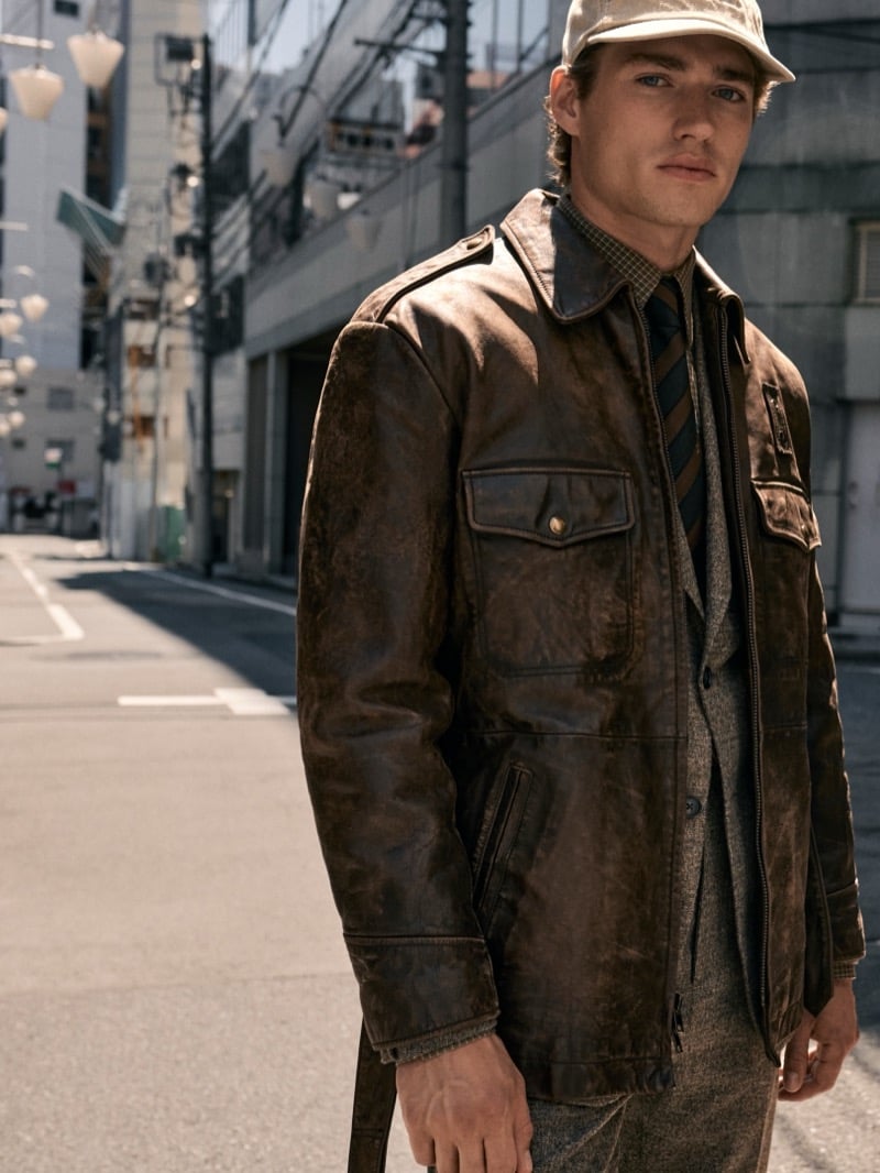 Valentin Humbroich wears a distressed brown leather jacket over a gray suit with a striped tie and a pale cap on a Tokyo street for Banana Republic's September 2026 edit