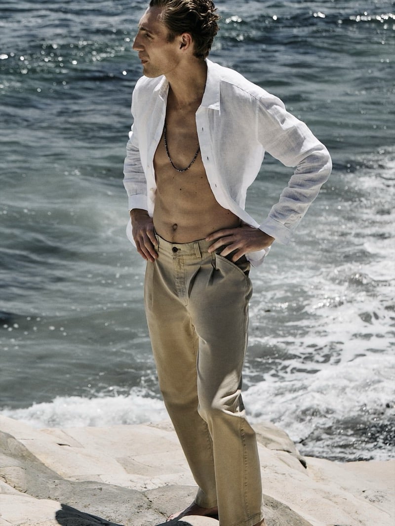 Aubrey O'Mahony on coastal rocks in an open standard fit linen shirt and trousers with a beaded necklace