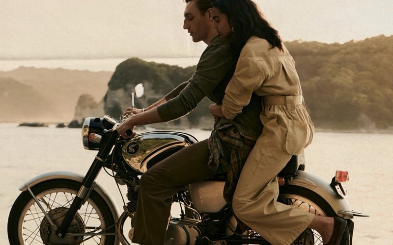 Aubrey O'Mahony rides a vintage Honda motorcycle along the coast with a woman in a tan trench coat behind him