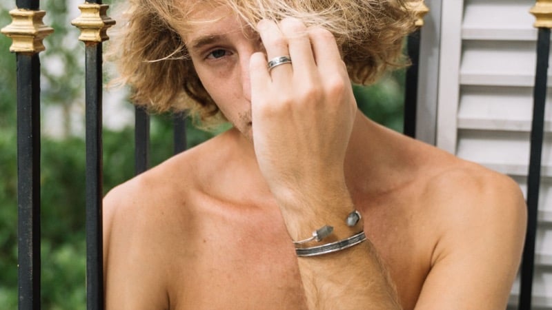 A shirtless man on a balcony wearing a beaded cuff bracelet, hand in his hair