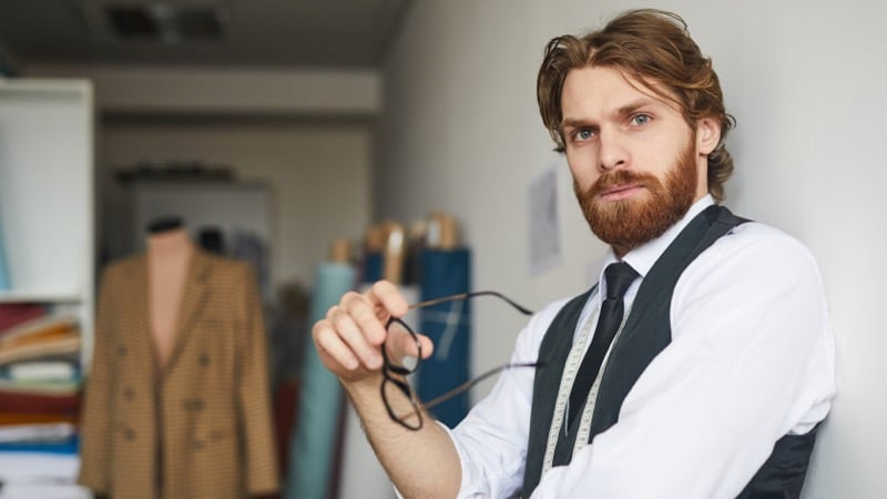 A bearded man in a vest and tie leans against a wall in a tailoring studio, holding a pair of glasses, with a jacket on a mannequin and fabric bolts behind him