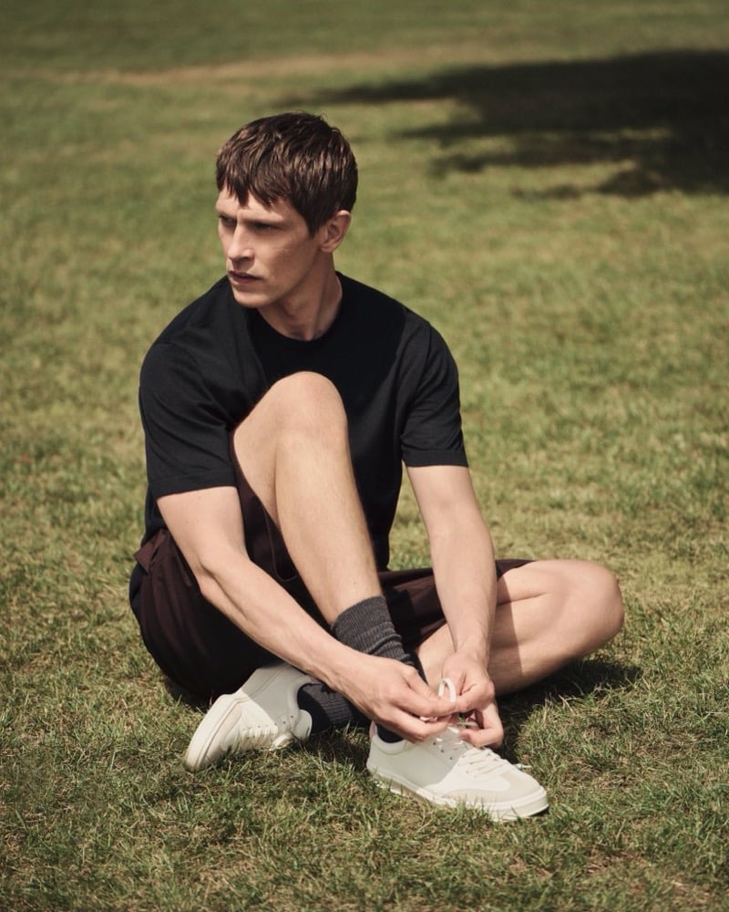 Mathias Lauridsen sits on the grass in a black crewneck t-shirt and brown shorts, tying the laces on a pair of white leather sneakers, for the BOSS by Beckham summer capsule.