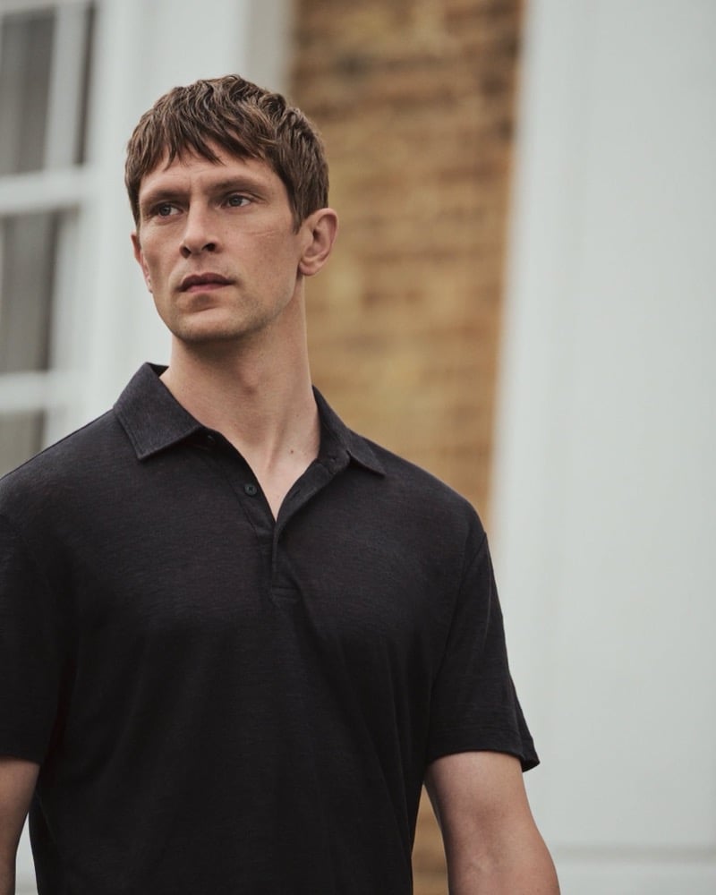 Close-up of Mathias Lauridsen in a black linen polo shirt, looking off to the side against a brick and white building facade, for the BOSS by Beckham summer capsule.