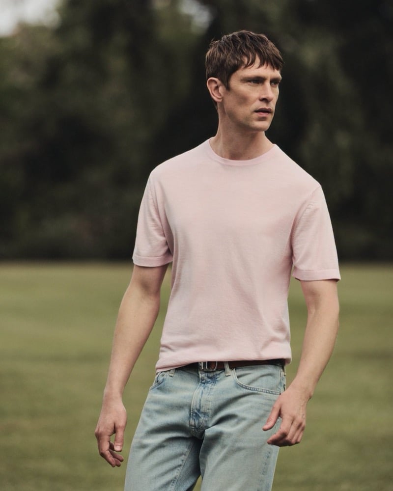 Mathias Lauridsen walks across a grass lawn in a pale pink crewneck t-shirt and light-wash denim jeans with a brown leather belt, for the BOSS by Beckham summer capsule.