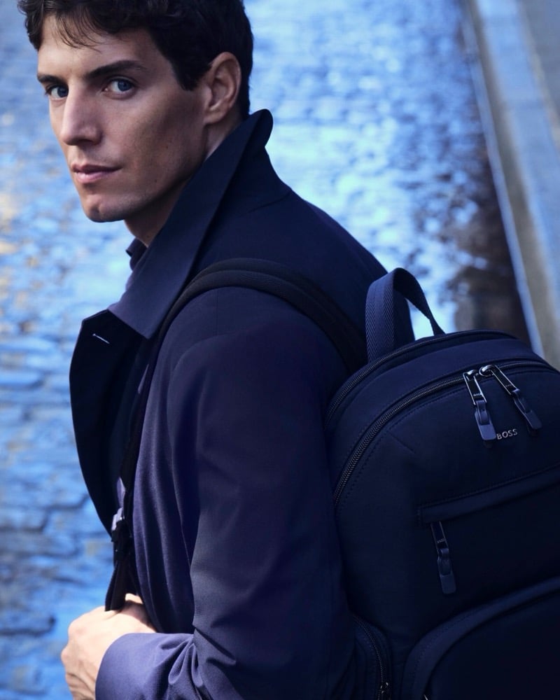 Marcos Fecchino glances over his shoulder in a navy suit jacket and BOSS backpack on a wet cobblestone street for the BOSS Performance edit.