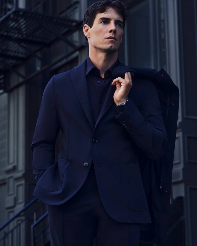 Marcos Fecchino stands in a navy suit with a matching jacket draped over one shoulder in front of a New York fire escape for the BOSS Performance edit.