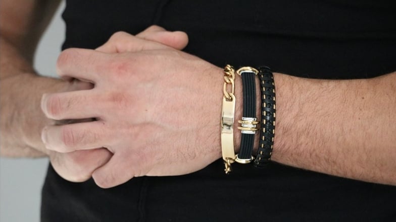 A man's wrist stacked with a gold chain bracelet, a rubber band, and a woven leather bracelet