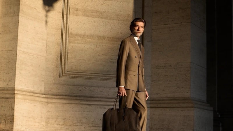 Patrick Petitjean walks past a sunlit travertine wall in a tobacco double-breasted suit, holding a leather tote bag, for Brioni's fall-winter 2026 campaign