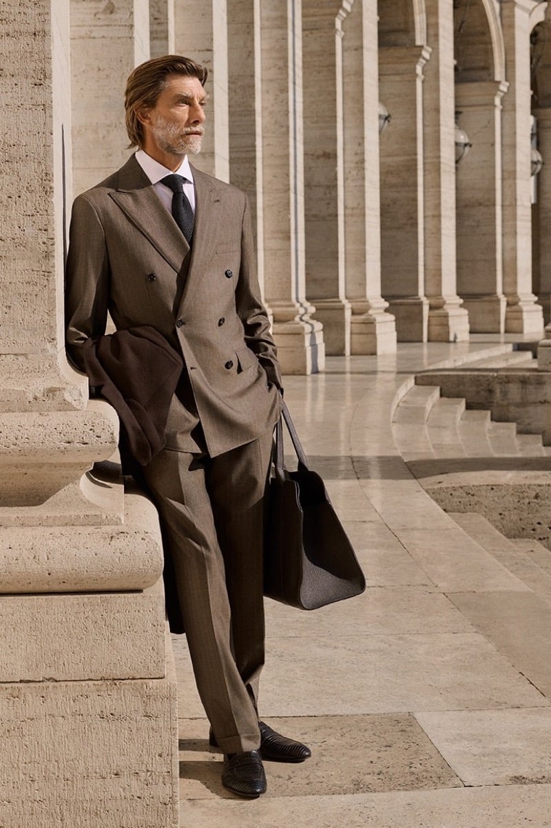 Patrick Petitjean leans against a travertine column in a pinstriped double-breasted suit, a topcoat over his arm and a leather tote in hand, for Brioni's fall-winter 2026 campaign