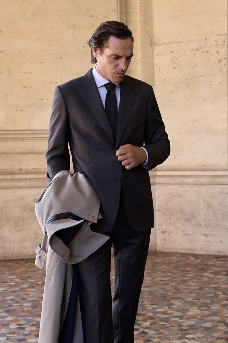 Jerome Adamoli buttons a charcoal single-breasted suit while carrying a topcoat over his arm in a marble-floored Roman colonnade for Brioni's fall-winter 2026 campaign