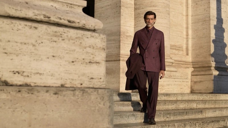 Patrick Petitjean descends travertine steps in a burgundy double-breasted suit with a coat folded over one arm for Brioni's fall-winter 2026 campaign