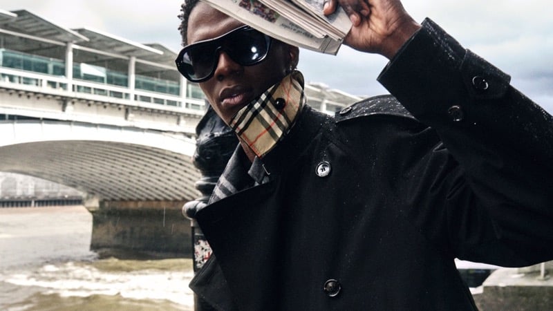 Cherif Douamba in a black trench coat holding a newspaper overhead in the rain, with Burberry check lining showing at the collar