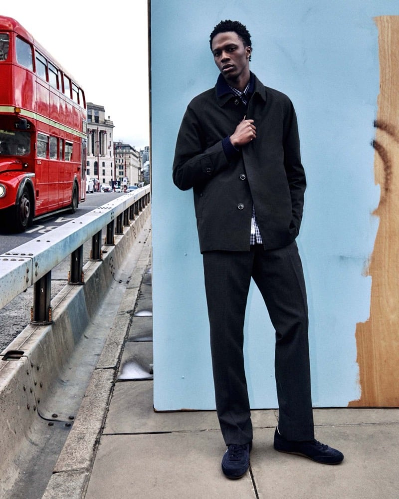 Cherif Douamba in a dark car coat over a checked shirt standing beside a red double-decker bus in London