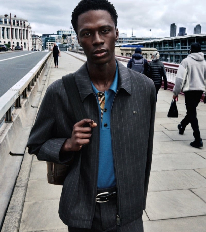 Cherif Douamba in a gray pinstripe Harrington jacket over a teal polo shirt with Burberry check trim, crossing a bridge in London