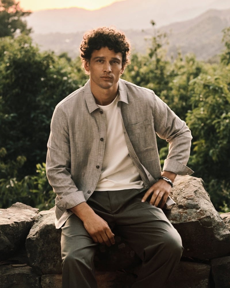 Simon Nessman wears a stone-gray overshirt over a white tee with olive trousers seated on a stone wall at sunset for Calvin Klein