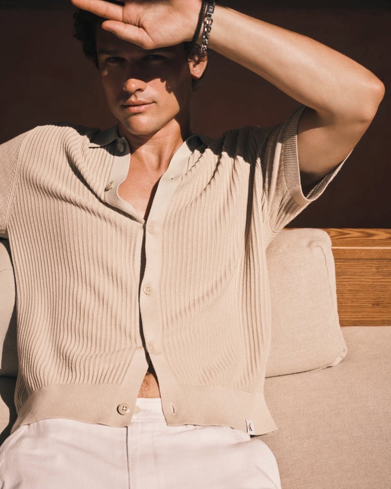 Simon Nessman wears a ribbed tan knit button-down with white trousers, shielding his eyes with a stacked-bracelet arm on a linen couch for Calvin Klein