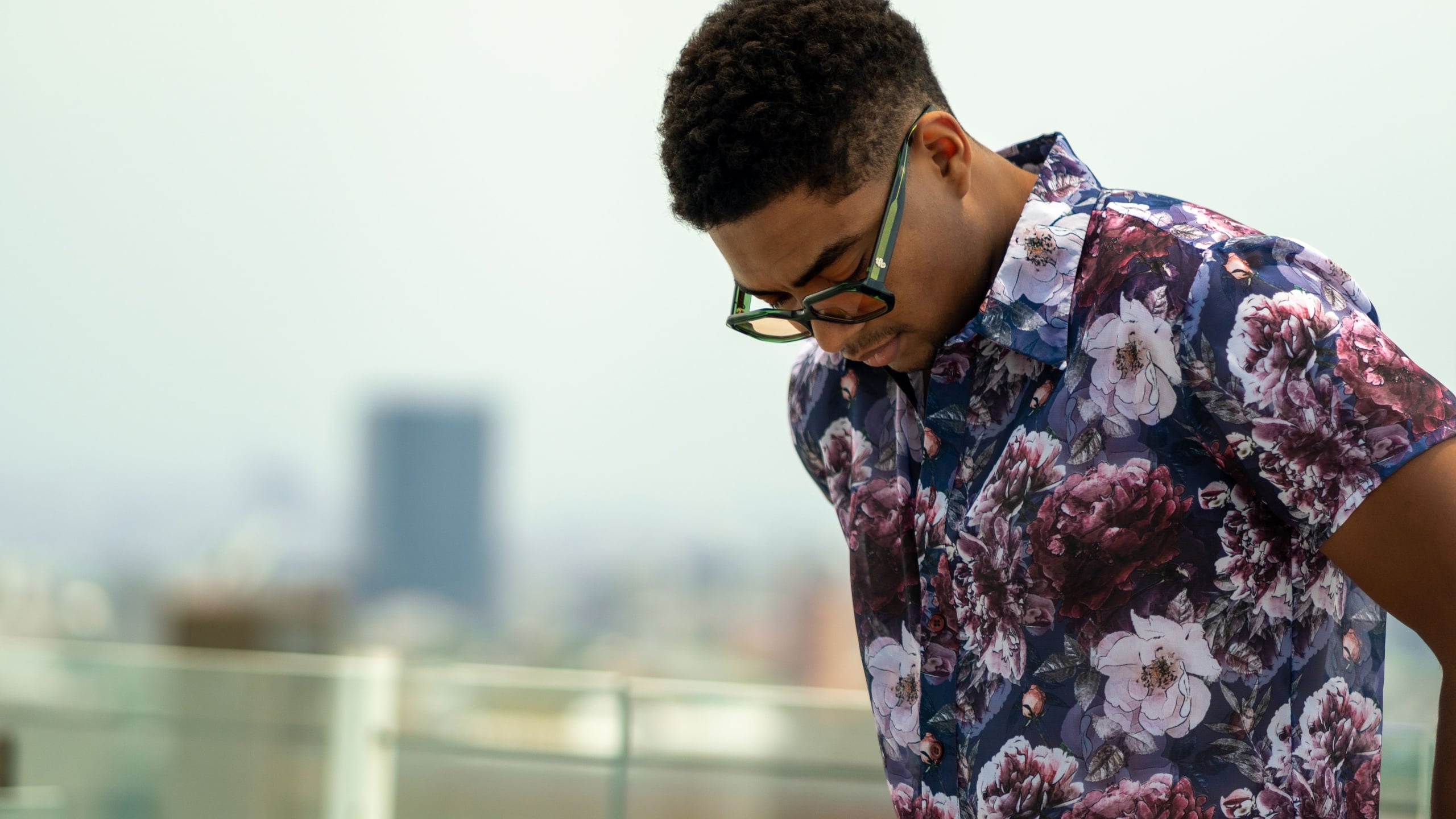 A man in sunglasses wears an open floral camp collar shirt with burgundy and white blooms on a rooftop