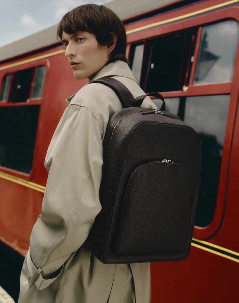 Erik Kettschick wears a beige trench coat and carries a dark leather backpack in front of a vintage red railway carriage for Carl Friedrik's fall 2026 campaign