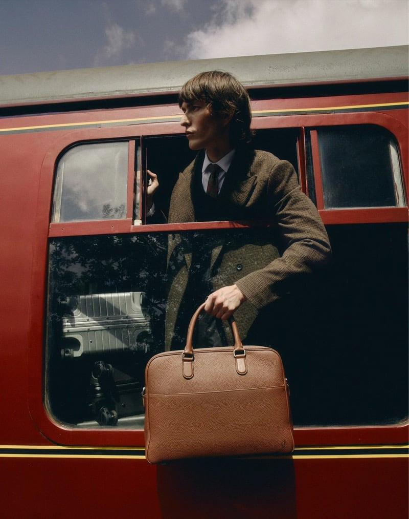 Erik Kettschik leans out of a train window holding a cognac leather briefcase, a silver hard-shell suitcase visible inside the carriage, for Carl Friedrik's fall 2026 campaign
