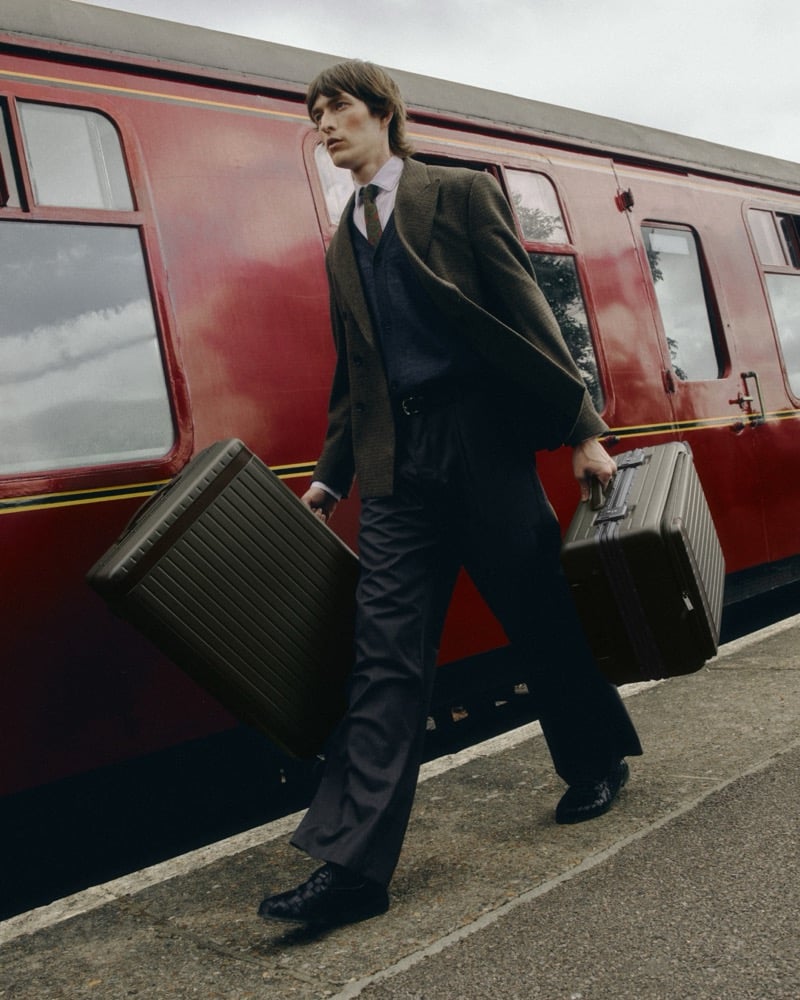 Erik Kettschik walks along a railway platform carrying two hard-shell suitcases beside a vintage red railway carriage for Carl Friedrik's fall 2026 campaign